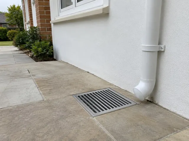 Outdoor metal drain grate beside a white house wall and paved path in Wahroonga