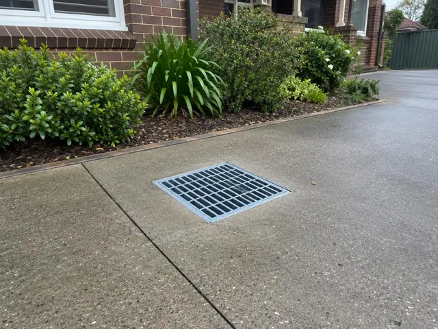 A clean stormwater grate set in a concrete driveway beside garden beds in Waitara